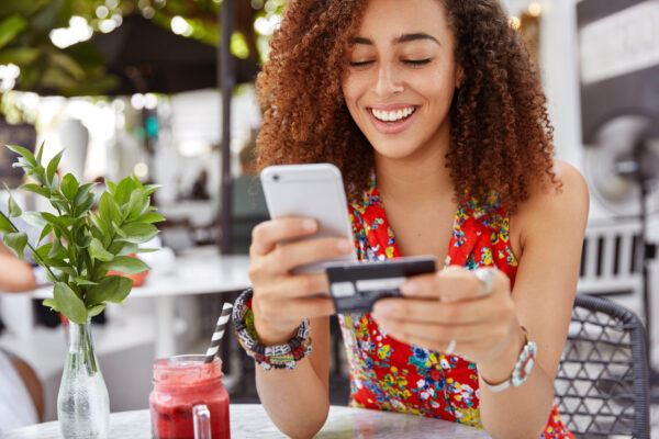 Young female with cheerful expression, holds smart phone and credit card.